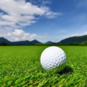 A golf ball resting on a perfectly manicured patch of green grass under a clear blue sky with a few fluffy white clouds.